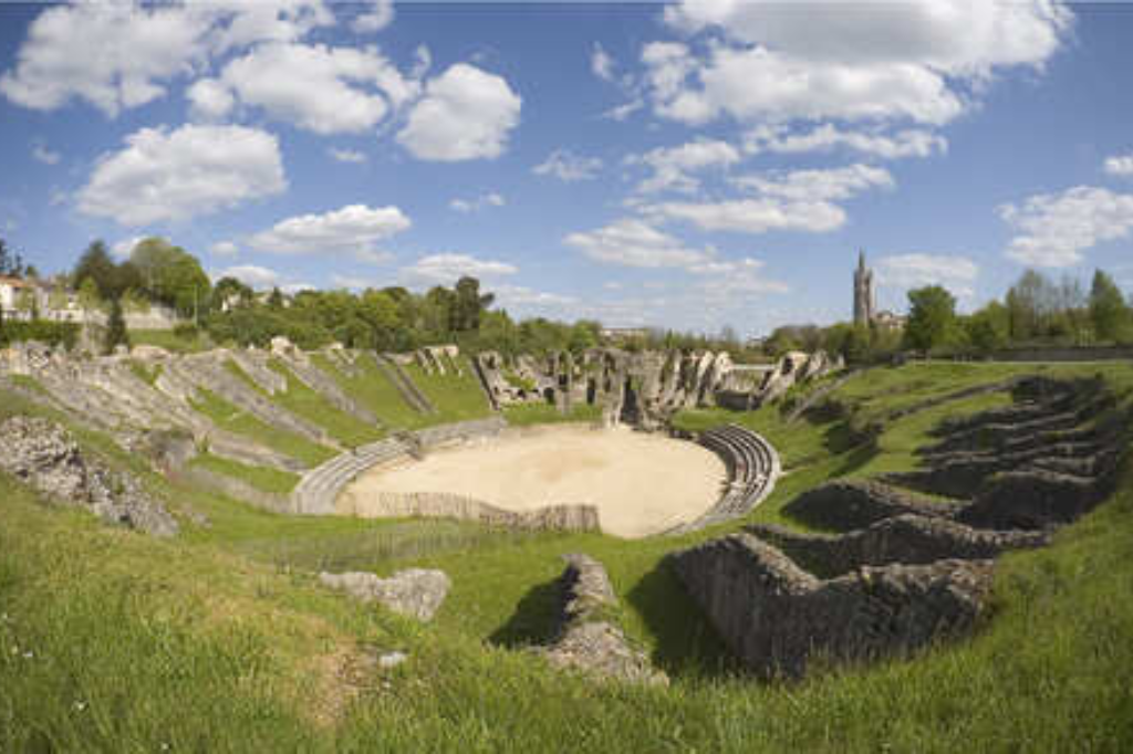 Vue panoramique des vestiges de l'amphithéâtre romain de Saintes, montrant l'arène centrale entourée de gradins recouverts d'herbe et de structures en pierre, sous un vaste ciel bleu parsemé de nuages blancs.