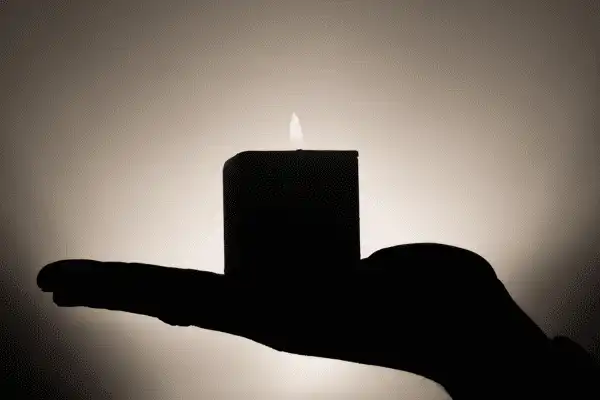 Silhouette en ombre chinoise d'une main ouverte portant une bougie carrée allumée, avec la lueur de la flamme se diffusant sur un fond clair et épuré.