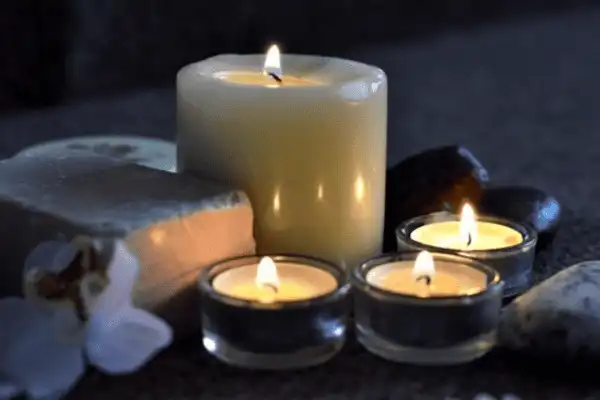 Composition d'ambiance spa au Domaine d'Angély : une grande bougie pilier allumée entourée de trois bougies chauffe-plat allumées, de galets de massage sombres et d'un pain de savon artisanal, avec une fleur blanche délicate sur fond sombre.