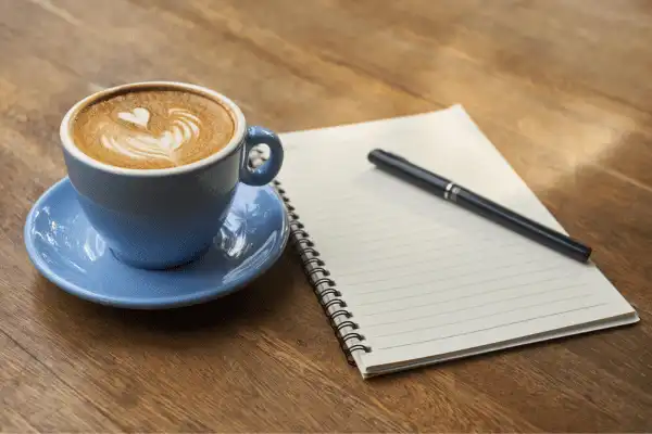 asse de café latte avec un motif de cœur dans la mousse, servie dans une tasse bleu ciel avec sa soucoupe, posée sur une table en bois clair à côté d'un carnet de notes et d'un stylo.