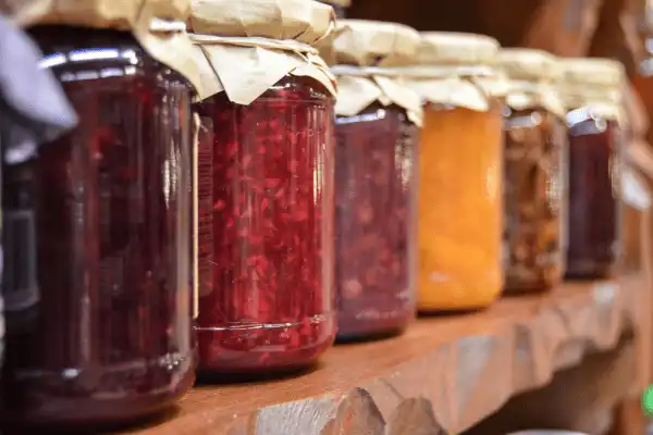 Alignement de bocaux de confitures artisanales aux couleurs variées (fraise, abricot, fruits rouges) avec couvercles recouverts de papier kraft, présentés sur une étagère en bois rustique.