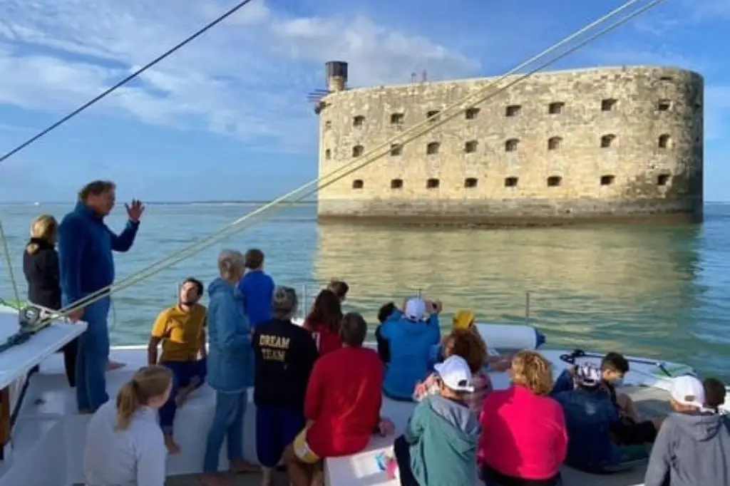 Un groupe de touristes à bord d'un catamaran s'approche du Fort Boyard en mer, sous un ciel bleu dégagé. Le fort en pierre se dresse majestueusement au-dessus de l'eau.