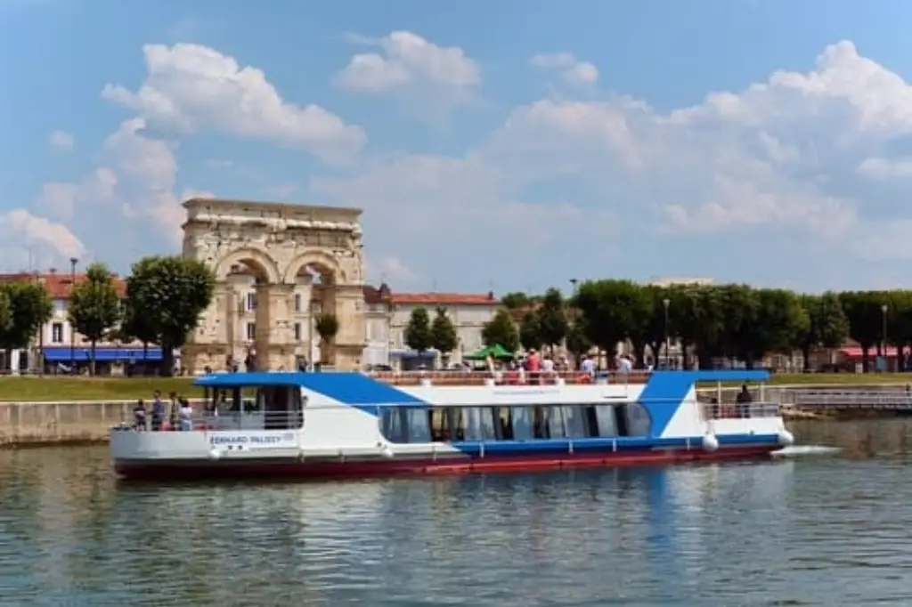 Un bateau de croisière blanc naviguant paisiblement sur les eaux calmes de la Charente, bordée d'arbres verdoyants et de roseaux sous un ciel lumineux.