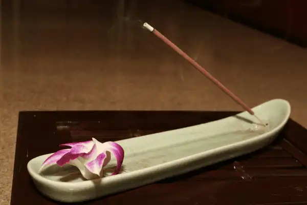 Un bâton d'encens allumé dégageant une fine fumée, posé sur un porte-encens en céramique verte avec une fleur d'orchidée rose et blanche délicate, sur une table sombre.