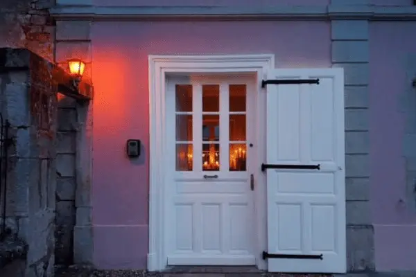 Entrée du gîte romantique au Domaine d'Angély : porte blanche éclairée de l'intérieur par des bougies, boîte à clés sécurisée pour arrivée autonome, mur rose poudré et lanterne rouge à la tombée de la nuit.