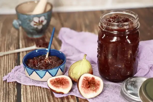 Figues fraîches coupées en deux et pot de confiture de figues maison présentés sur un linge violet avec une tasse de thé en arrière-plan.