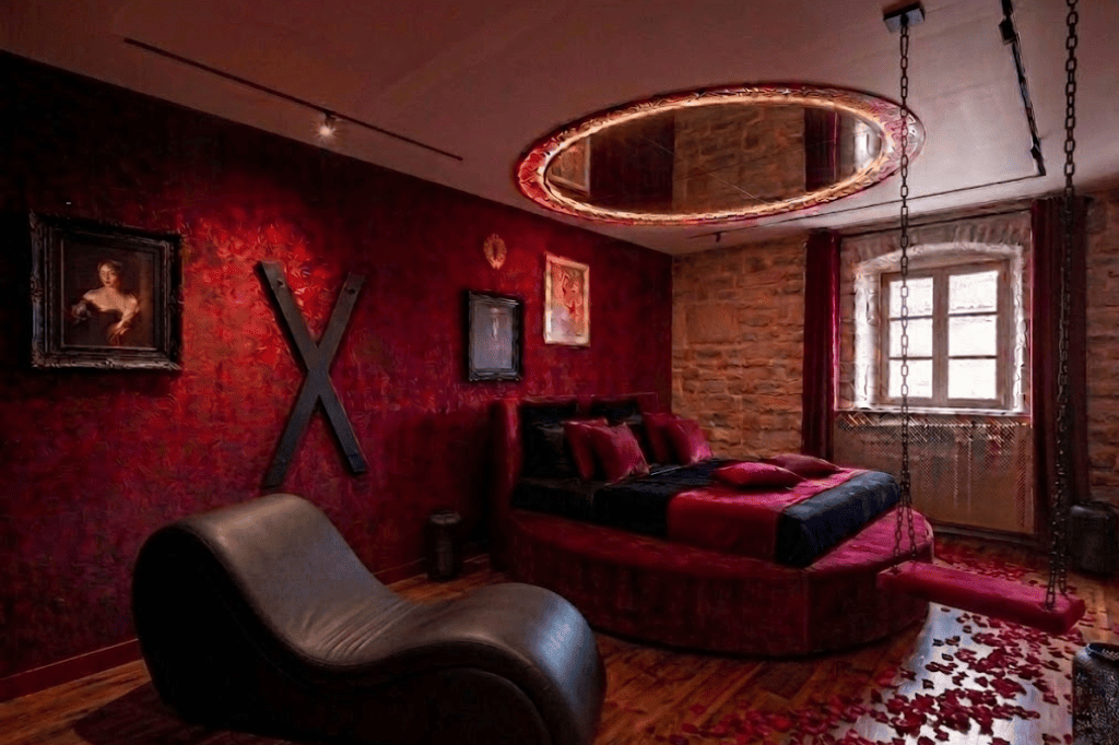 Love Room d'exception au Domaine d'Angély : grand lit rond en velours rouge, miroir ovale au plafond avec éclairage LED, murs texturés bordeaux avec portraits baroques encadrés ainsi qu'une croix de saint andré, mur en pierre apparente, miroir psyché, fauteuil tantra en cuir et sol parsemé de pétales de roses rouges.