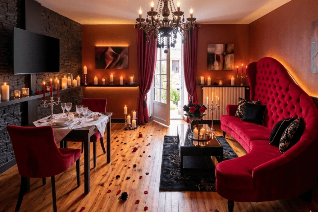 Rez-de-chaussée du gîte romantique au Domaine d'Angély : salon avec grand canapé en velours rouge, table dressée pour un dîner aux chandelles, pétales de roses sur parquet, lustre à pampilles et mur en pierre noire.