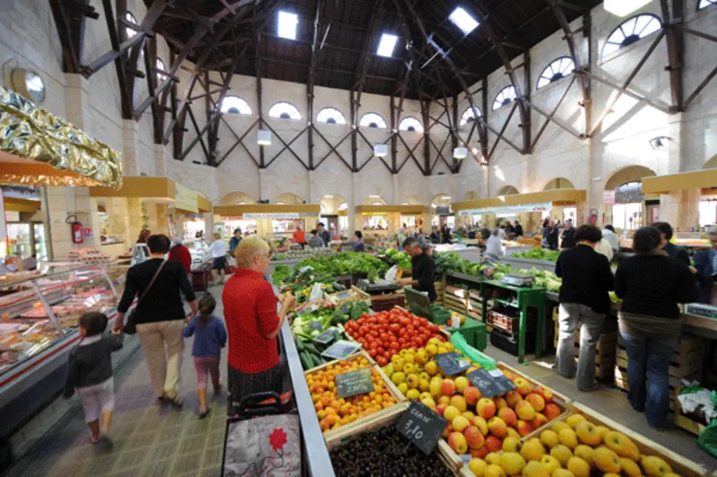 Intérieur animé de la Halle St Pierre à Saint-Jean-d'Angély, montrant une charpente monumentale en bois, des étals colorés remplis de fruits et légumes frais, et des clients faisant leurs courses.