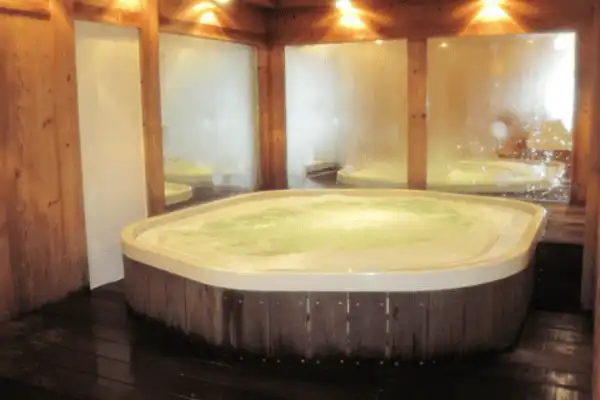 Jacuzzi installé dans une pièce intime aux murs boisés, avec une eau bouillonnante éclairée et des spots tamisés créant une ambiance chaleureuse.