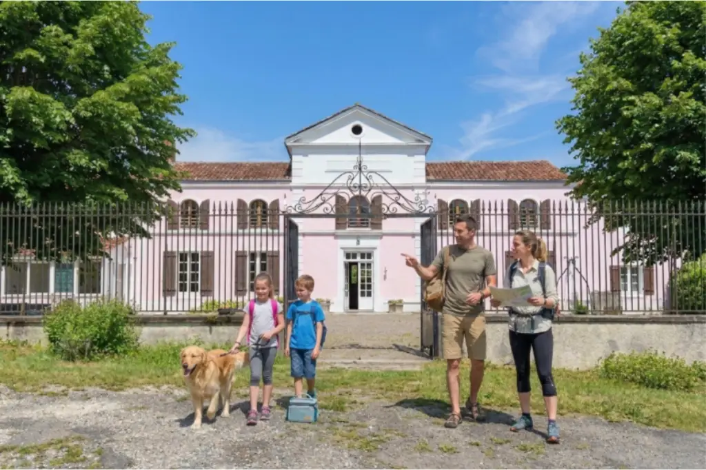 Une famille avec deux enfants et un Golden Retriever se tenant devant la grande maison rose et blanche du Domaine d'Angély. Les parents tiennent une carte de randonnée et le père montre une direction, sous un ciel bleu radieux.