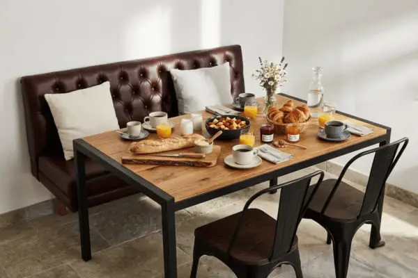 Table de petit-déjeuner copieux au Domaine d'Angély avec viennoiseries fraîches, baguette, jus d'orange, confitures artisanales et café, servie sur une table en bois massif avec banquette en cuir.