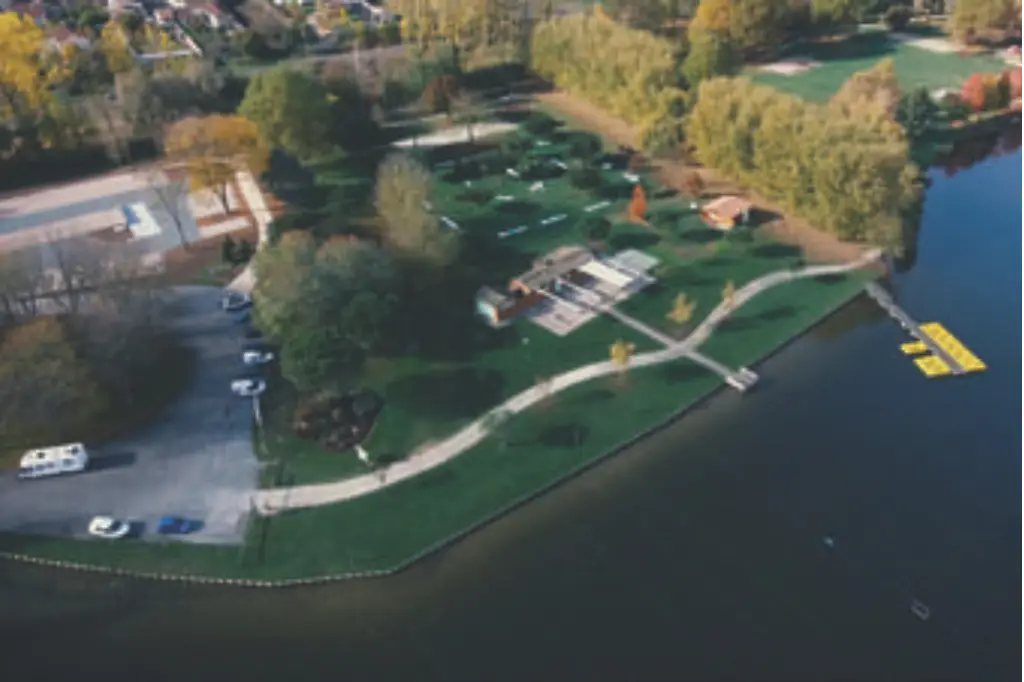 Vue aérienne du plan d'eau de Bernouet montrant les berges aménagées avec des sentiers de promenade, des zones de pelouse verdoyantes, des arbres en couleurs automnales et un ponton sur l'eau.