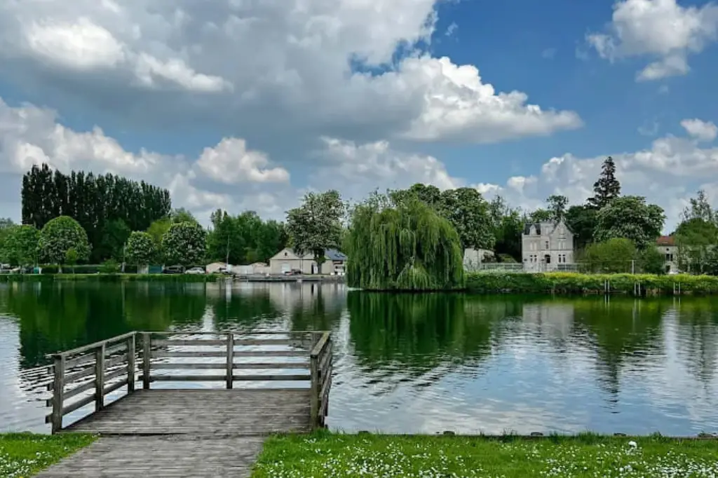 Un ponton en bois sur les bords de la Boutonne à Saint-Jean-d'Angély, avec des arbres verdoyants et une belle demeure ancienne se reflétant dans l'eau paisible sous un ciel nuageux.