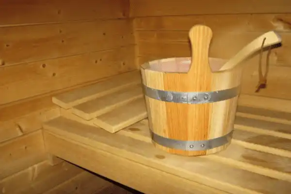 Seau en bois avec cerclage métallique et louche assortie posés sur les bancs d'un sauna en bois clair, prêt pour le rituel de la vapeur.