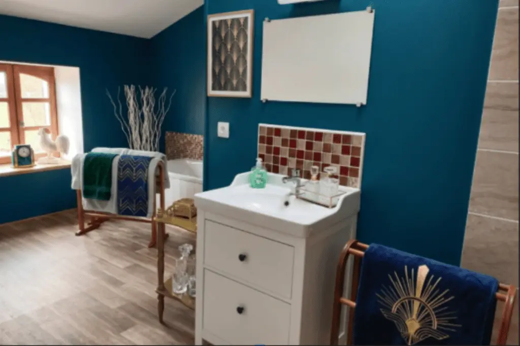Salle de bains de la suite Art Déco au Domaine d'Angély avec murs bleu canard, meuble vasque blanc, baignoire, douche à l'italienne et linge de maison aux motifs dorés.