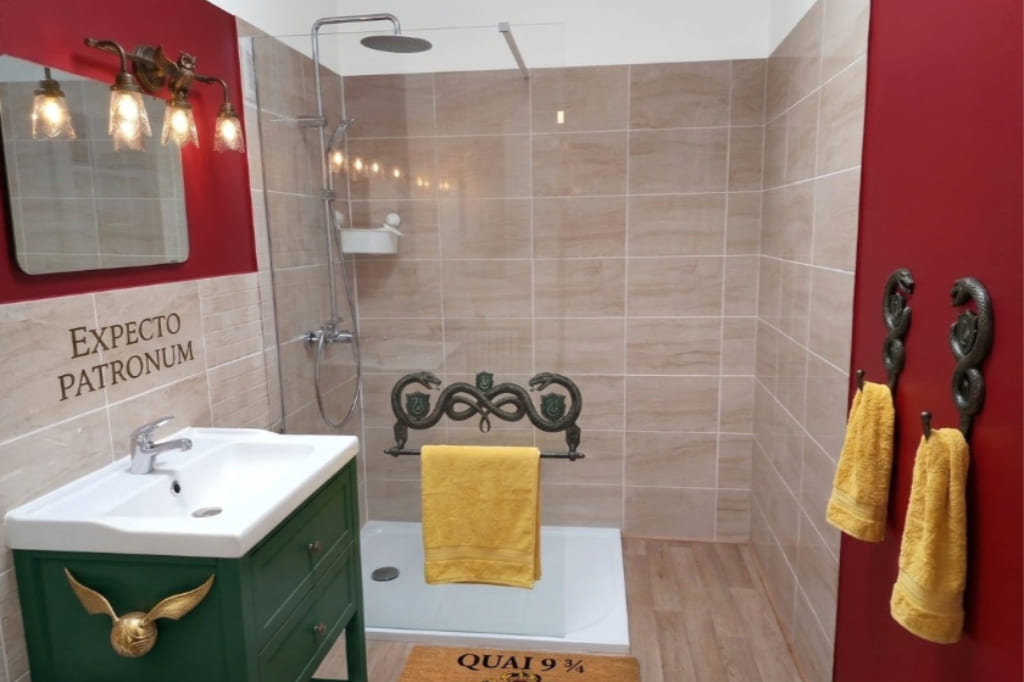 Salle de bains thématique Harry Potter au Domaine d'Angély avec douche à l'italienne, meuble vasque vert avec Vif d'Or sculpté, murs rouge bordeaux, inscription Expecto Patronum et tapis de bain Quai 9 ¾.