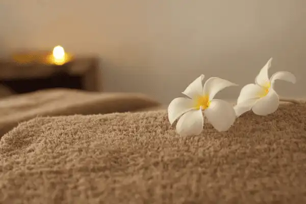 Gros plan sur une serviette de bain épaisse et moelleuse de couleur beige, ornée de deux fleurs de frangipanier blanches et d'une bougie allumée en arrière-plan flou.