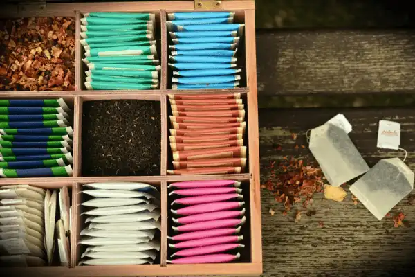 Vue de dessus d'un coffret en bois compartimenté contenant une large variété de sachets de thé colorés et de thé en vrac, posé sur une table en bois rustique.