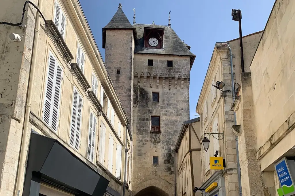 Vue en contre-plongée de la Tour de l'Horloge à Saint-Jean-d'Angély, une tour médiévale en pierre avec des mâchicoulis, un toit en ardoise pointu et une horloge intégrée, encadrée par des bâtiments urbains sous un ciel bleu.