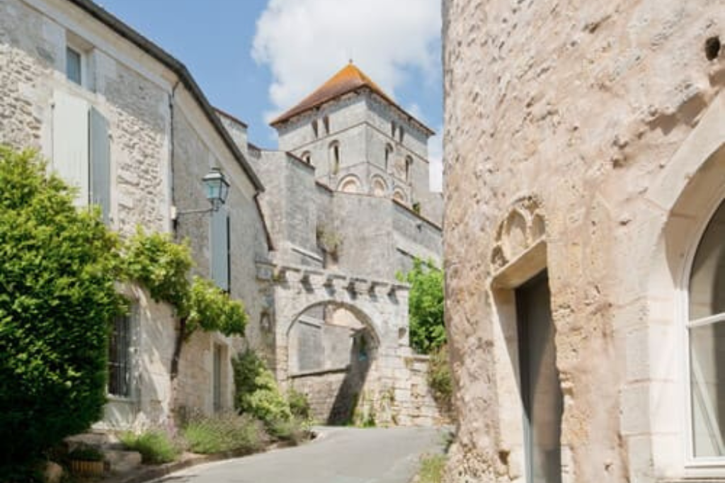 Rue montante du village médiéval de Saint-Sauvant avec ses maisons en pierre calcaire, une arche ancienne traversant la ruelle et l'église romane dominant l'horizon sous un ciel bleu.