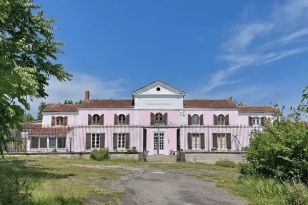Vue de la façade principale du Domaine d'Angély, une grande demeure de maître rose aux volets marron, située derrière une grille en fer forgé sous un ciel bleu radieux.
