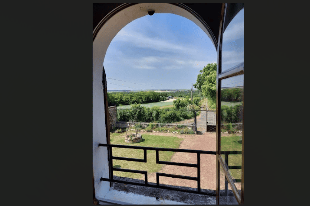 Vue dégagée depuis une fenêtre à arche de la suite Viking sur la cour avant verdoyante, un puits ancien, et les champs de la campagne environnante au Domaine d'Angély.