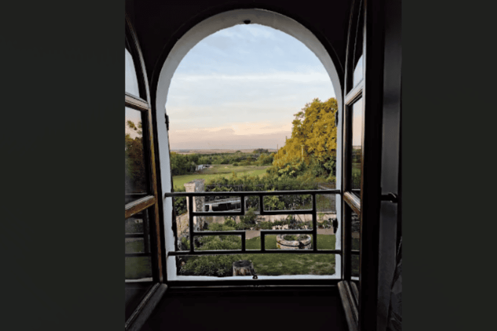 Vue panoramique depuis la fenêtre à double arche du salon Harry Potter sur le jardin arboré, le puits ancien et l'horizon champêtre du Domaine d'Angély au coucher du soleil.